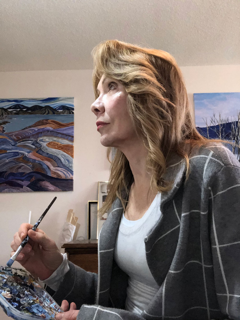 Liz Nicholson at work in her studio, holding a brush and palette, two of her paintings on the wall behind her